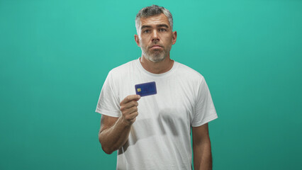 Fototapeta premium Man holds blue creditcard between fingers in a studio teal backdrop, white tshirt visible, hand raised and lips pursed showing uncertainty; financial concern.