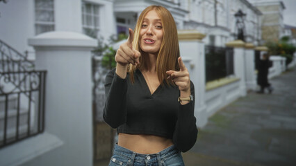 Fototapeta premium Young redhead woman pointing both index fingers at camera, smiling and showing bare stomach on a residential street; playful.