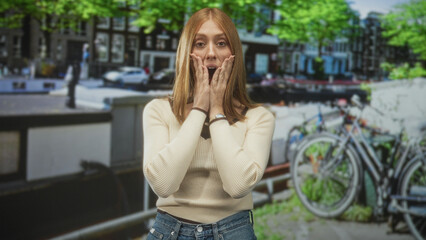 Fototapeta premium Woman touching hands to face with surprised expression, beige sweater and jeans on street near canal and bicycles; surprise curiosity.