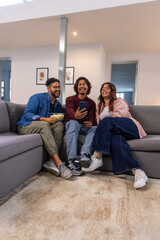 Naklejka premium Indian friends sitting on gray sofa at home in sneakers, sharing smartphone and bowl of popcorn