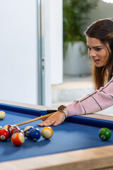 Fototapeta premium Woman leaning over blue pool table at home aiming pool shot in pink sweater, copy space