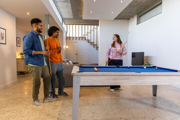 Diverse friends playing pool around blue felt table in modern home rec room, holding drinks