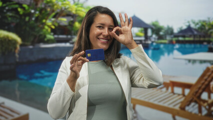 Hispanic middle age brunette woman holds blue creditcard and makes ok sign with right hand, smiling...