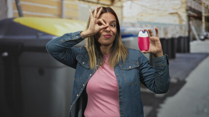 Fototapeta premium Middle aged hispanic woman holds pink toothpaste bottle near face, makes ok sign to eye and smiles in airport baggage claim carousel area; playful confidence.