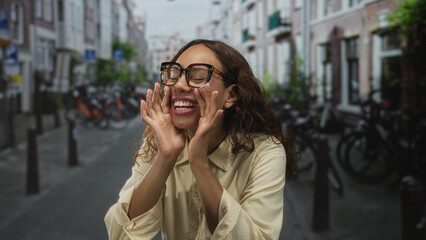 Fototapeta premium Woman with hands cupped around mouth and glasses on street lined with bicycles and narrow buildings; joyful playfulness.
