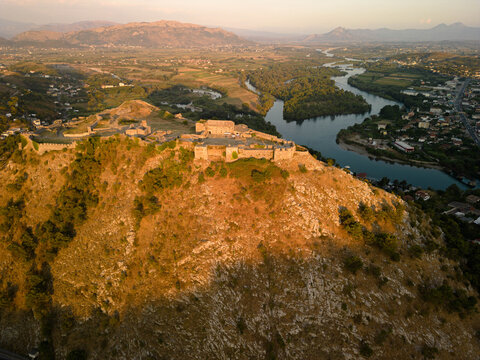 Aerial view of Rozafa Castle overlooking rivers and valley near Shkoder at sunset, golden hour light illuminating historic fortress and Albanian countryside landscape