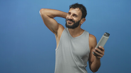 Fototapeta premium Man holding shampoo bottle, hand behind neck showing bare arm in blue studio; self care grooming confidence.