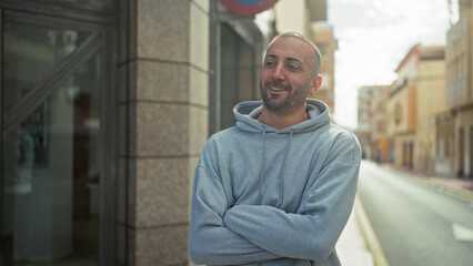 Obraz premium Man with arms crossed smiling, face visible, wearing grey hoodie beside stone building on a street; urban calm.
