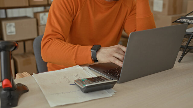 Man holds head and writes on invoice with pen beside laptop and calculator amid stacked cardboard boxes and barcode scanner in building; stress finances.