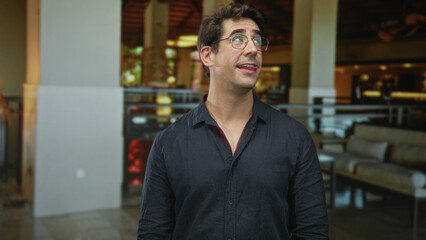 Fototapeta premium Man in dark shirt wearing glasses, smiling and sticking out tongue while looking up by lounge couches and railing in building; playful curiosity.