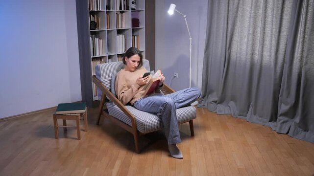 Young woman in a cozy living room sits in an armchair with a book while checking her smartphone, balancing traditional reading and digital media during a relaxed evening routine