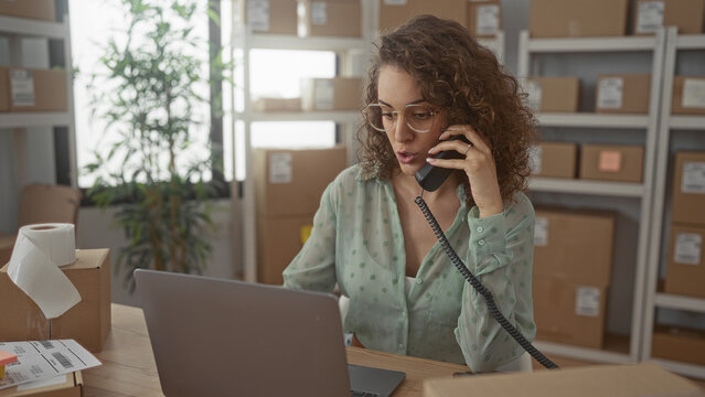 Woman talking on corded phone holding handset while working at laptop amid shipping boxes in office building; productive small business.