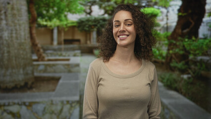 Fototapeta premium Woman smiling at camera with curly hair and visible neck, standing on a stone park walkway beside a low stone building and trees, wearing a beige sweater; warmth connection happiness.