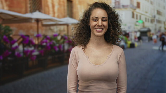 Woman in pink top showing cleavage standing on a restaurant terrace by umbrellas and purple flowers, turning head to the right on a cobbled street; quiet contemplation.