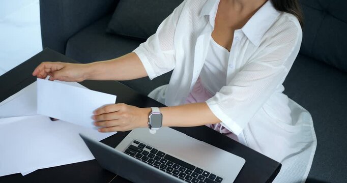 Female reviews bills and statements. Woman sorts financial documents at home desk. Woman diligently reviews and sorts financial documents on her living room table under soft daylight light
