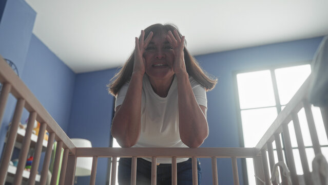 Woman leaning over crib covering her face with hands in a nursery inside a building; gentle playfulness.