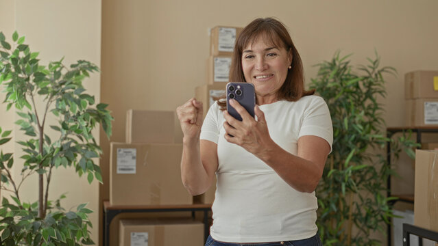 Woman raising fist and holding smartphone beside stacked cardboard boxes in a building; small business triumph.
