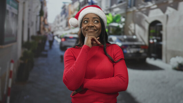 Woman wearing santa hat and red sweater with hand on chin and arms crossed on a snowy city street; festive joy.