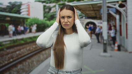 Obraz premium Young hispanic woman covering ears on crowded train station platform, wearing white top and jeans, tracks and commuters visible; distress hearing pain.
