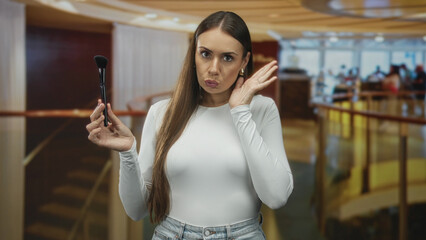 Fototapeta premium Woman holding makeup brush with hand to face, covering mouth and touching cheek in building; surprise uncertainty.
