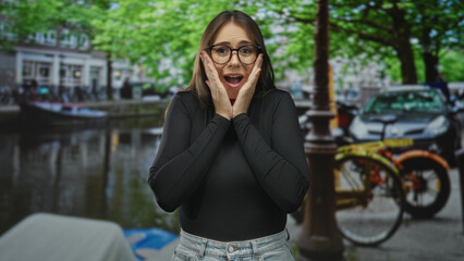 Fototapeta premium Young woman touching hands to cheeks in surprise on street beside a canal with parked bicycle and cars; surprise wonder.