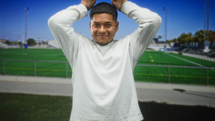 Man with beard and hands on head smiling showing teeth in white long sleeve shirt at a stadium building, soccer field visible  joy casual. © Krakenimages.com