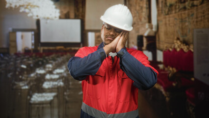 Fototapeta premium Man construction worker in hard hat presses hands to cheek for sleep gesture in building; tired rest.