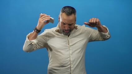Fototapeta premium Middle age man with beard holding a smartphone and raising his arm while dancing against a bright blue studio backdrop; joy celebration.