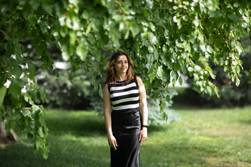 Fototapeta premium Horizontal portrait of a joyful young woman with warm reddish-brown hair, standing outdoors in natural daylight against a softly blurred green garden background