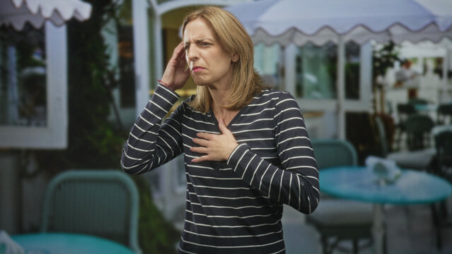 Woman touching ear then clutching chest in street cafe near outdoor tables and umbrellas, gingerly raising hands toward throat with pained expression; distress.