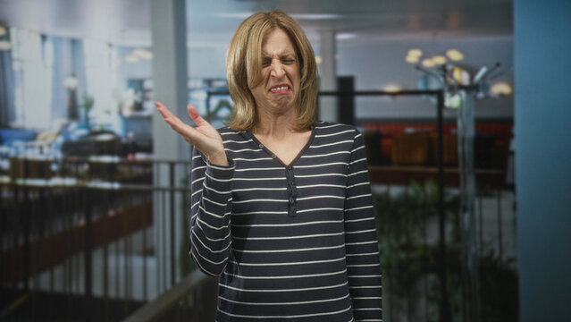 Woman covers nose with hand and recoils in a building atrium near railing and lamps while grimacing; disgust rejection.