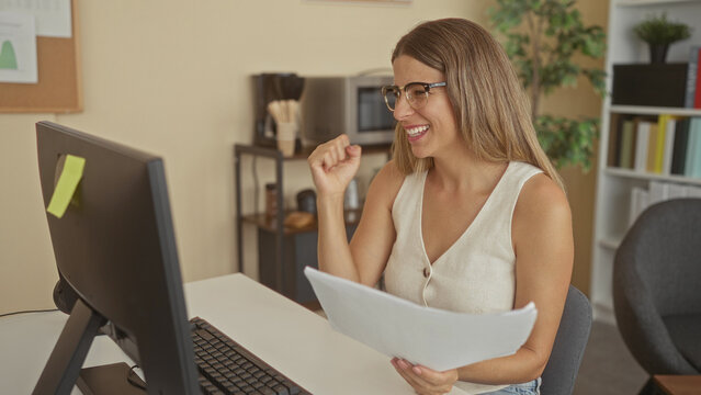 Woman wearing glasses typing at computer, holding papers and smiling with a small fist pump in building; remote work satisfaction.