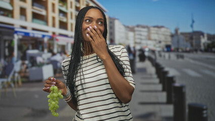 Fototapeta premium Woman holding green grapes and wearing striped shirt covers mouth with hand while standing by outdoor cafes and bollards on street; surprise delight.