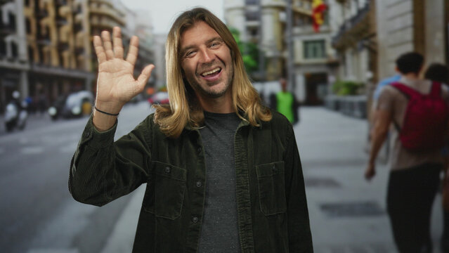 Blond man waves hand on busy urban street amid blurred pedestrians and vehicles in daytime; friendly welcome.