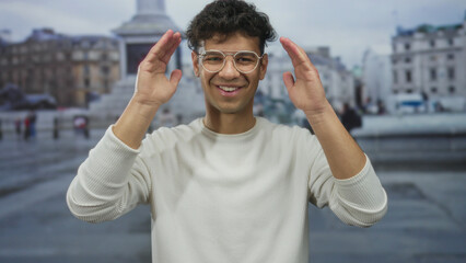 Obraz premium Young hispanic man wearing glasses smiles and covers his eyes with both hands on a wet city street; surprise.
