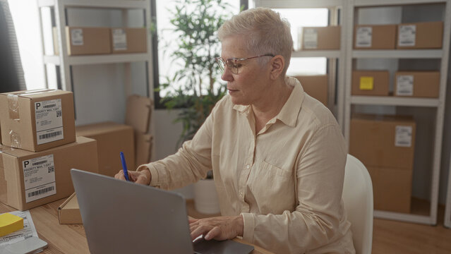 Woman packing parcels and typing on laptop at office building desk with stacked boxes and visible shipping labels; small business concentration.