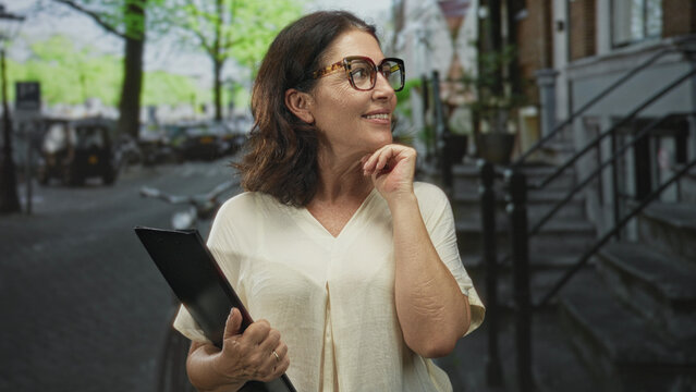 Woman holding folder touches chin on street, wearing glasses and beige blouse with bicycles and stoop visible; thoughtful optimism.