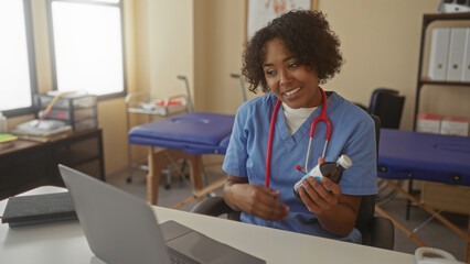 Fototapeta premium Young african american woman doctor holds medicine bottle beside laptop during online clinic consultation in building; compassion trust.