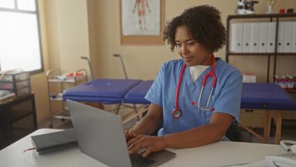 Young african american woman in blue scrubs typing on laptop at white desk in clinic office; serenity productivity.