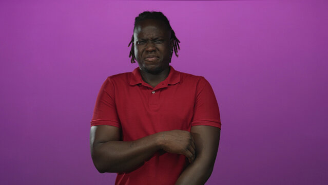 Young african american man grimacing while holding his arm in studio with purple backdrop; disgust reaction.
