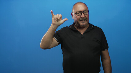 Fototapeta premium Man making hand sign with index and pinky raised and thumb extended in studio, wearing black polo and glasses; affection connection.