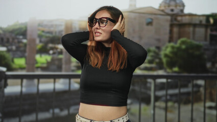Obraz premium Woman redhead holding hands over ears, wearing glasses on a building balcony by ancient ruins; discomfort isolation.