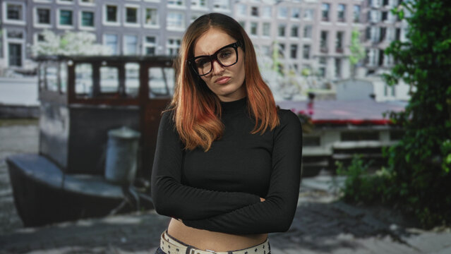 Woman wearing glasses with crossed arms and pursed lips by a moored boat on street, black crop top and belt visible; annoyance solitude.