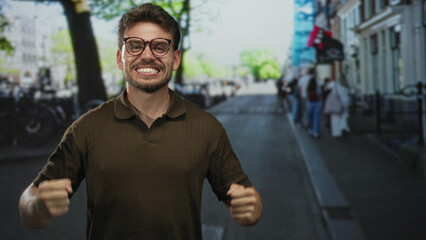 Fototapeta premium Man with clenched fists and raised arms on a busy city street, wearing glasses and smiling; triumph celebration success.