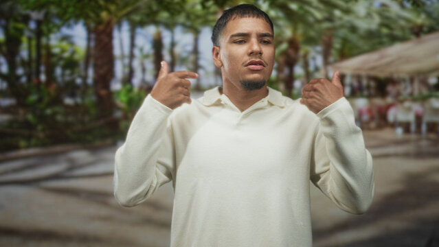 Man young hispanic adjusting shirt at chest with right hand, wearing cream polo in forest cafe by palm trees and canopy seating; contemplation.