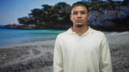 Young man wearing glasses stands facing camera at a beach backdrop in a studio lit by soft light  calm confidence. © Krakenimages.com