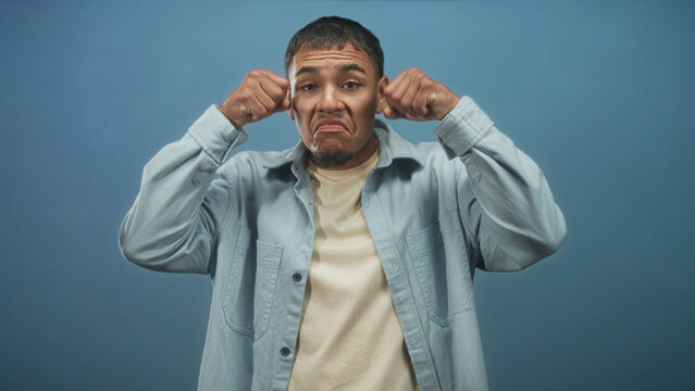 Man pulls at his eyes with clenched fists against a blue studio backdrop, miming exaggerated crying and mock frustration while wearing a light shirt; playful.