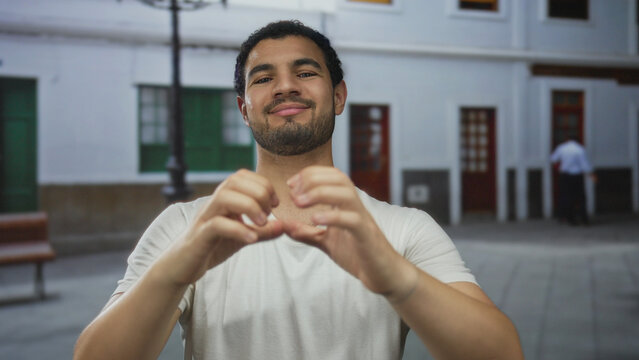 Young hispanic man in white tshirt smiling and spreading hands on sunlit busy street; warm welcome.
