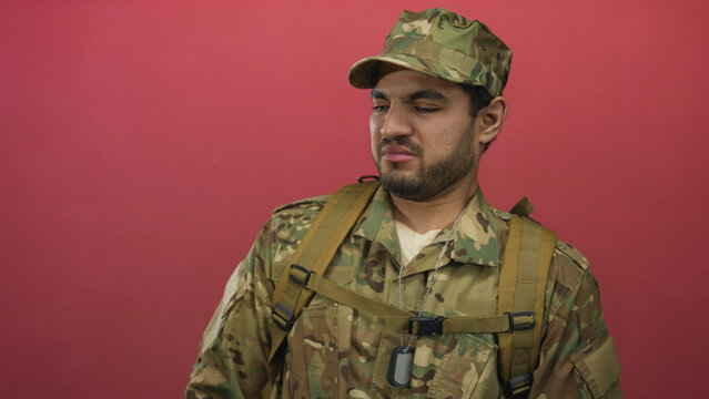 Man in camouflage uniform scrunches nose and frowns against a red studio wall under bright lighting; disgust.