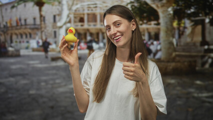 Fototapeta premium Woman smiling and holding a yellow rubber duck while giving thumbs up on a historic street with building facade; joyful play.